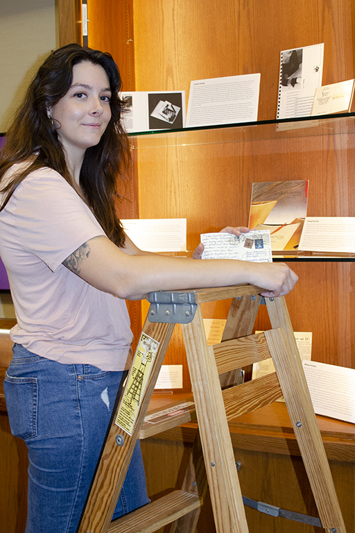 Photograph of graduate student Hannah Obsorn standing on a ladder in from of an exhibit case. 