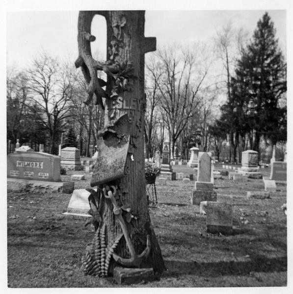 Black and white photograph of tombstone in the shape of a tree trunk with additional symbols of an anchor, ivy, and scroll included. 