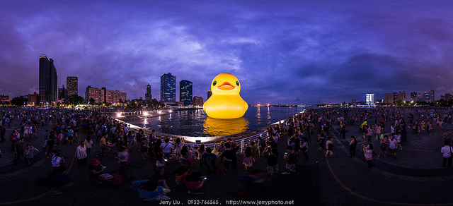 a giant rubber duck in a city harbor under a dramatic sky with a crowd looking on
