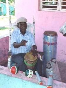 Picture of a man in a cap seated on a small outdoor patio. The patio is painted pink with blue accents. The man is holding a small drum between his knees. There are small percussion instruments in front of him and a large drum on his left-hand side.