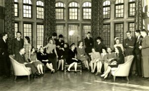  Black and white photograph of a group of students gathered together in front of large windows. 