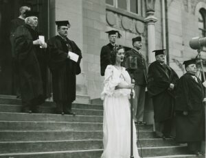  Black and white image of a woman in a white dressing leading a procession of men in graduation gowns. 
