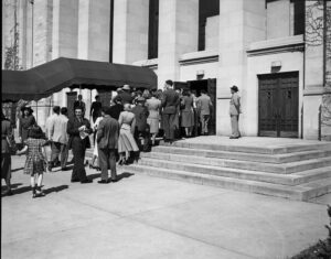  Black and white photograph of a crowd entering a building. 