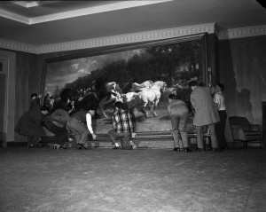  Black and white photograph of workers hanging a large painting on a wall. 