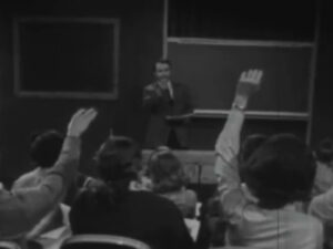  Still image from a black and white film showing a teacher calling on students with raised hands. 