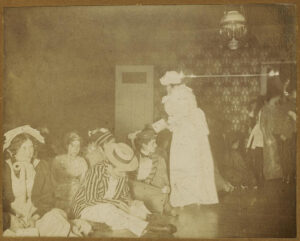 Women sitting on floor or standing, some dressed in men's clothing. 