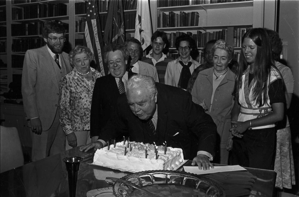 An older Herman B Wells blows out candles on his birthday cake in front of friends and staff