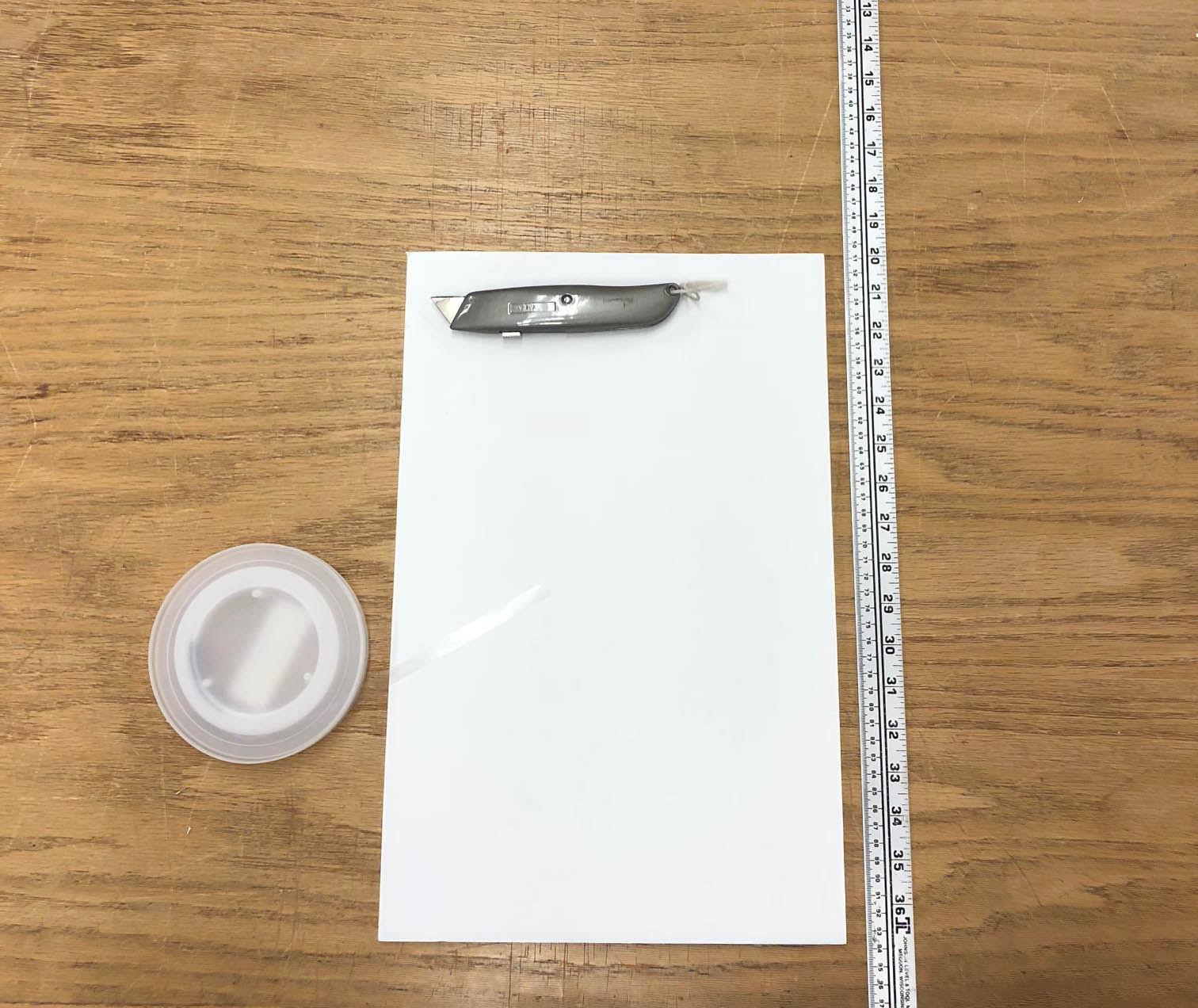 A roll of polyethylene book strapping, a piece of foam core, a utility knife, and a ruler on a table.