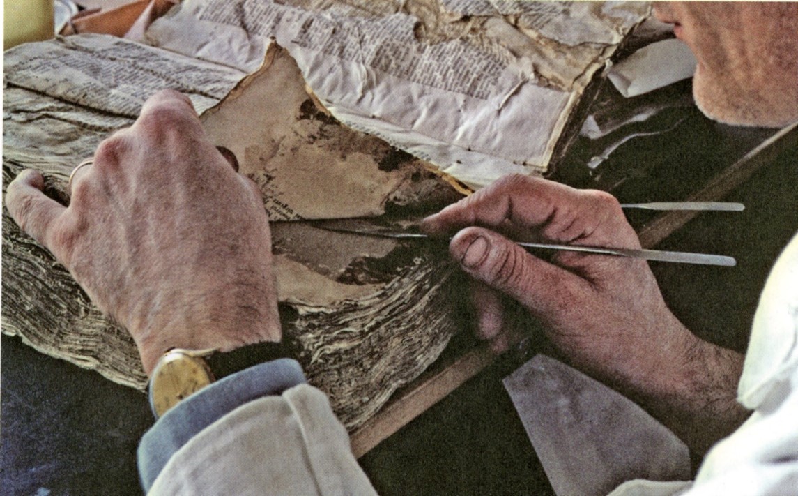 Color photograph of a man's hands using instruments to pull apart pages of a damaged book.