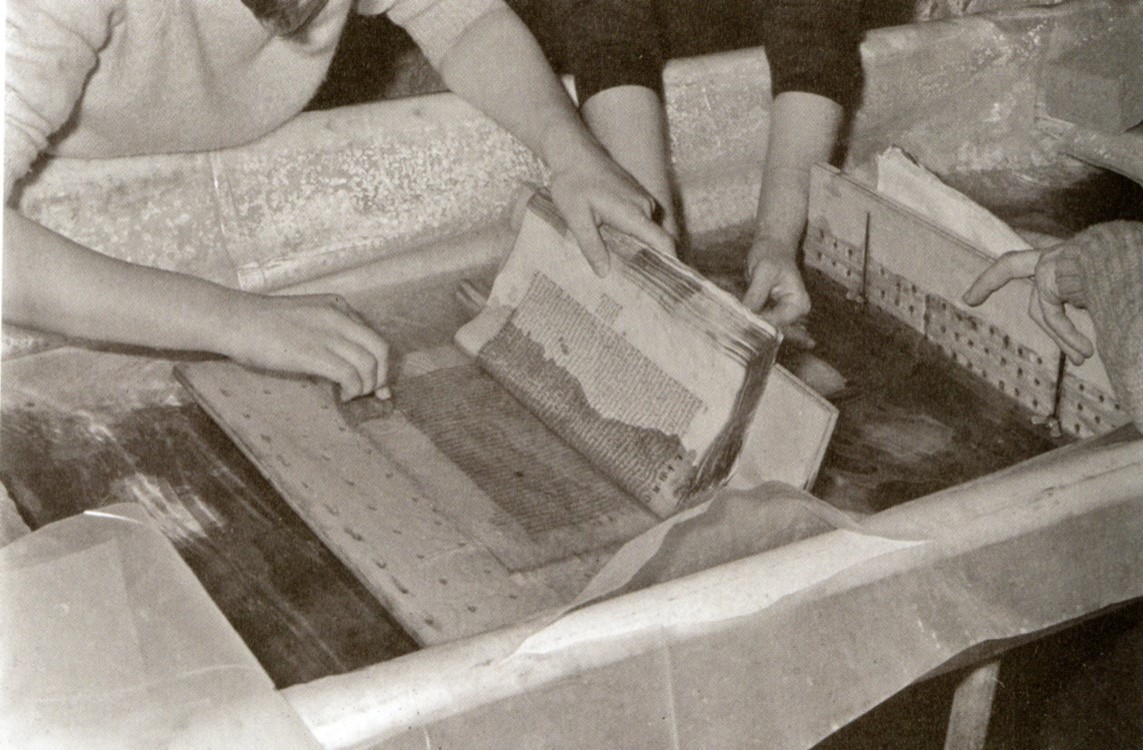 Black and white photograph of the arms of two women cleaning a damaged book. 