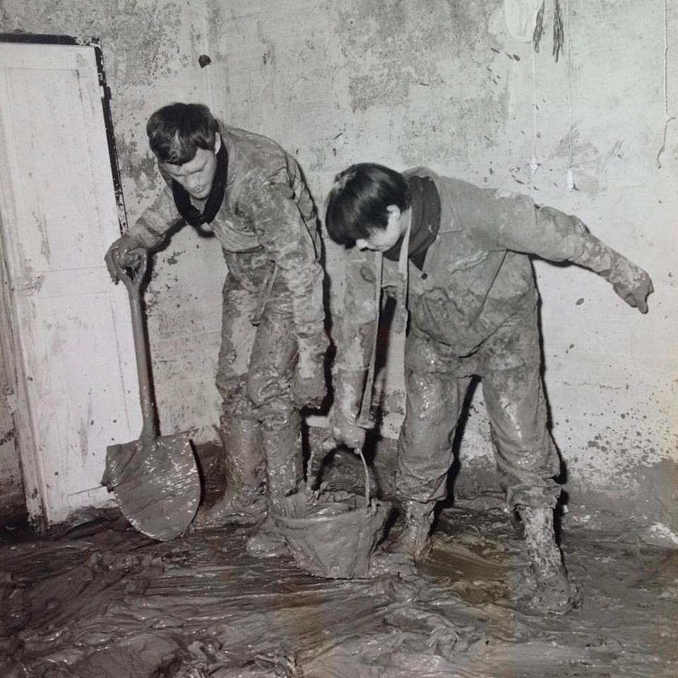 Black and white photograph of two workers with shovel and bucket cleaning up mud in a hallway.
