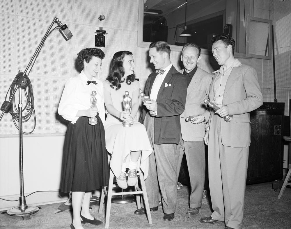This is a black and white photograph of a group of 5 individuals who are each holding their Oscar award. Three men wearing suits stand to the right, while 2 women one standing and the other seated on a stool are to the left. 
