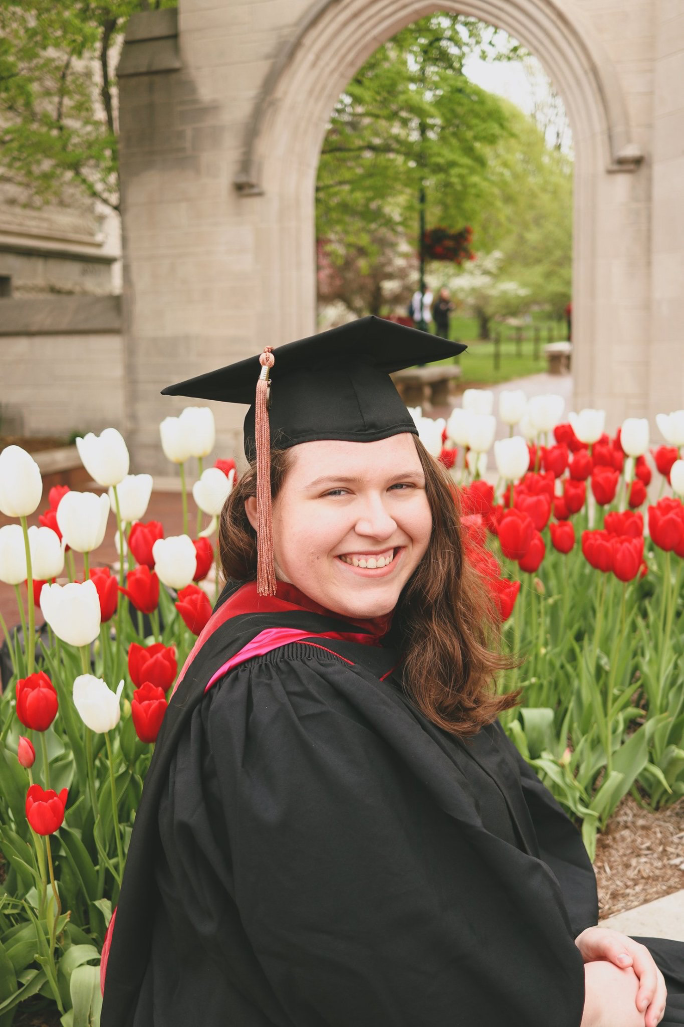This is a color photograph of a student in a graduation cap and gown. She is seated in the front of the archway of the IU Sample Gates and is surrounded by red and white tulips. 