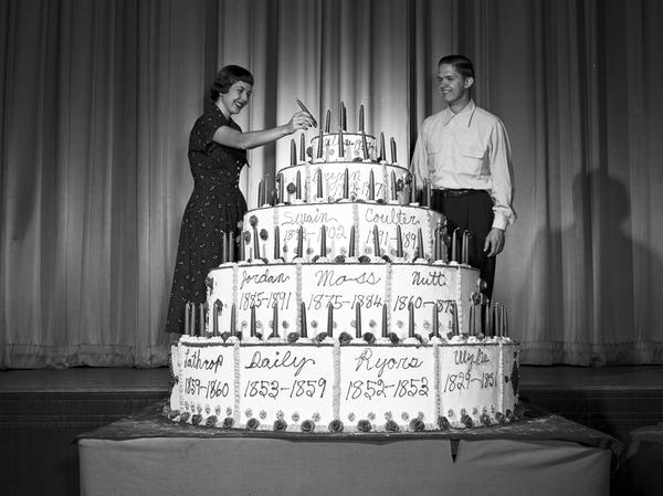 Black and white photograph of 2 students standing beside a large 5-tier cake with tapered candles. 
