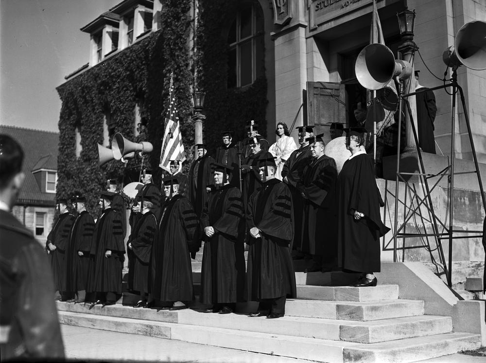 Black and white photograph of the Freshman Induction ceremony. Robed faculty and staff including President Wells stand on the front steps of the Student Building. 