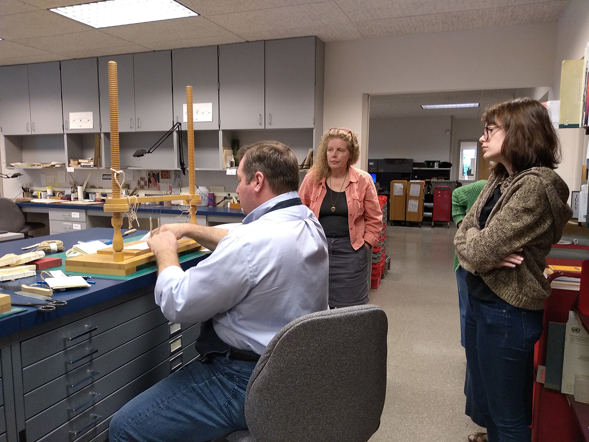 Conservation staff watching Andrew sewing signatures on to raised cords.