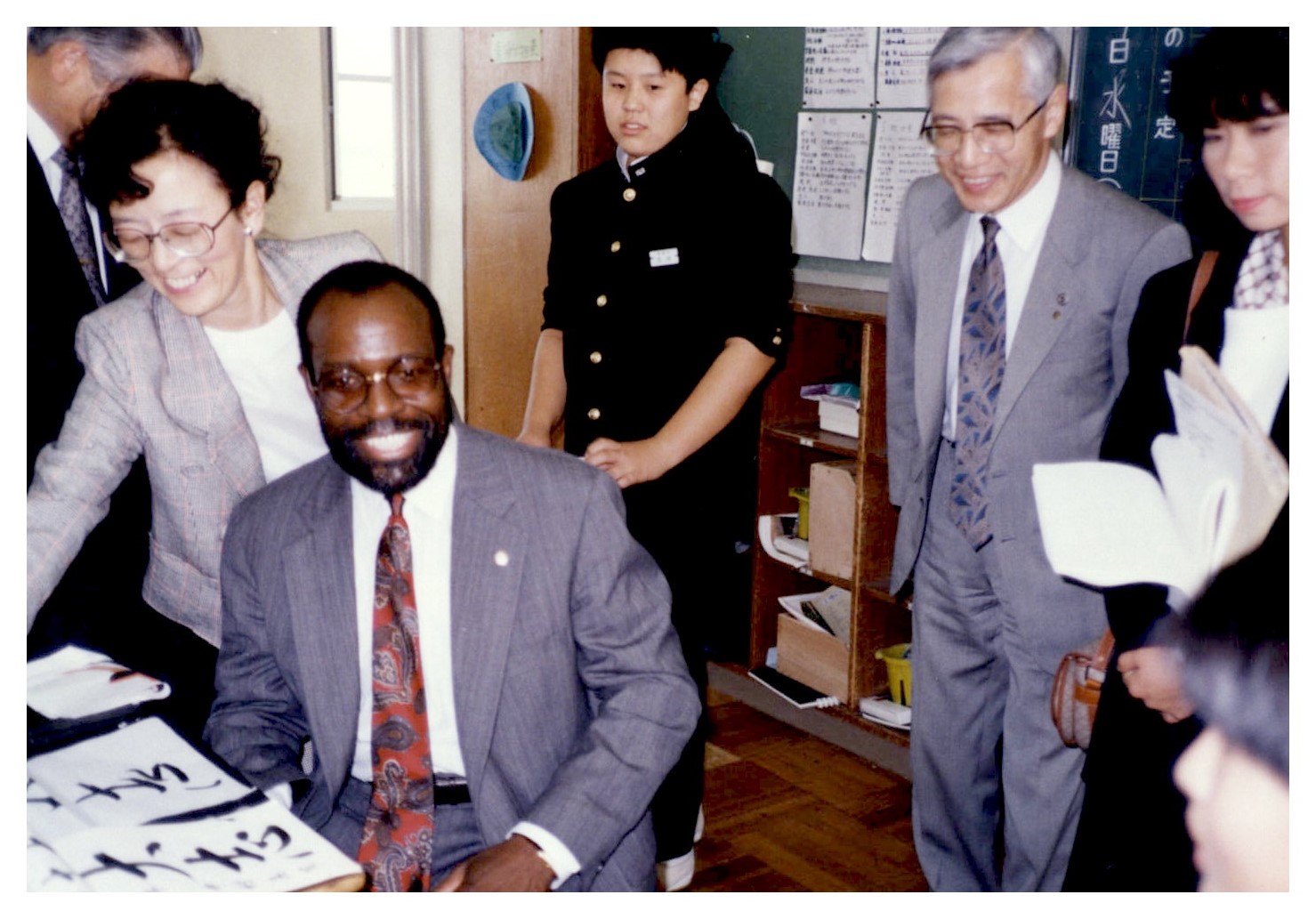 Charlie Nelms sits at a desk. Two women and two men stand nearby. On the desk are pieces of paper with Japanese character calligraphy.
