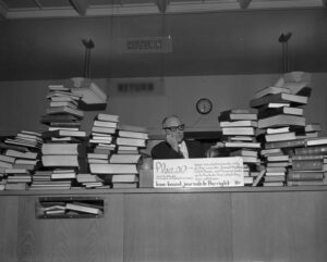Books piled high on the library circulation desk with a man resting his heand in his hand.