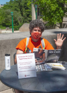 Greg Eismin, Circulation Supervisor in Wells Library, waves while sitting at a table outdoors reading the New Yorker magazine and wearing a face mask.