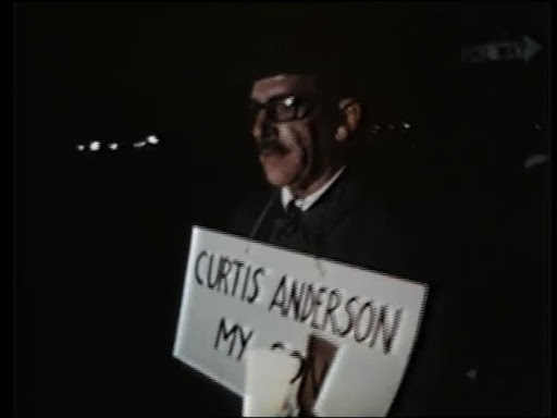 Man walking with a sign around his neck that says "Curtin Anderson, my son."