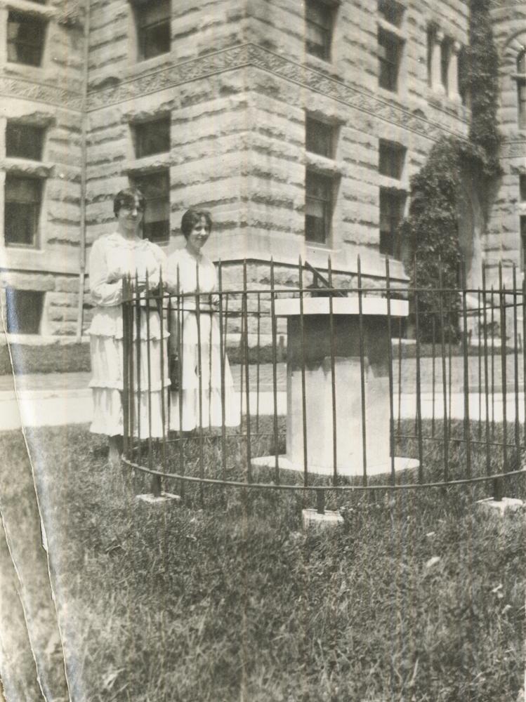 Photograph of 2 women in white dresses standing in front of a sundial.