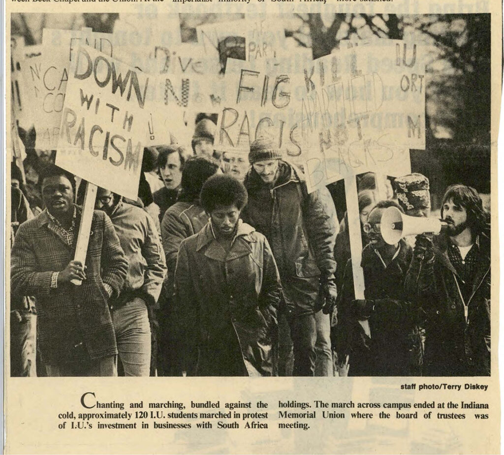 Protestors with signs and one with a bullhorn. Caption says, "Chanting and marching, bundled against the cold, approximately 120 I.U. students marched in protest of I.U.'s investment in businesses with South Africa holdings. The amrch across campus ended at the Indiana Memorial Union where the board of trustees was meeting."