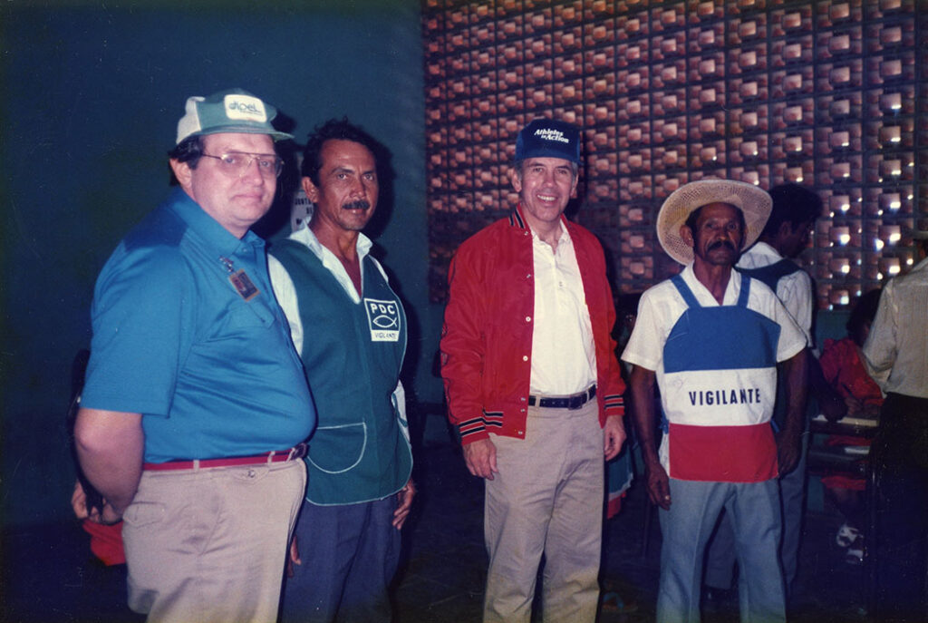 Photograph of four men, the third of them is Senator Richard Lugar. The man on the right is wearing an election observer vest with the label "Vigilante."