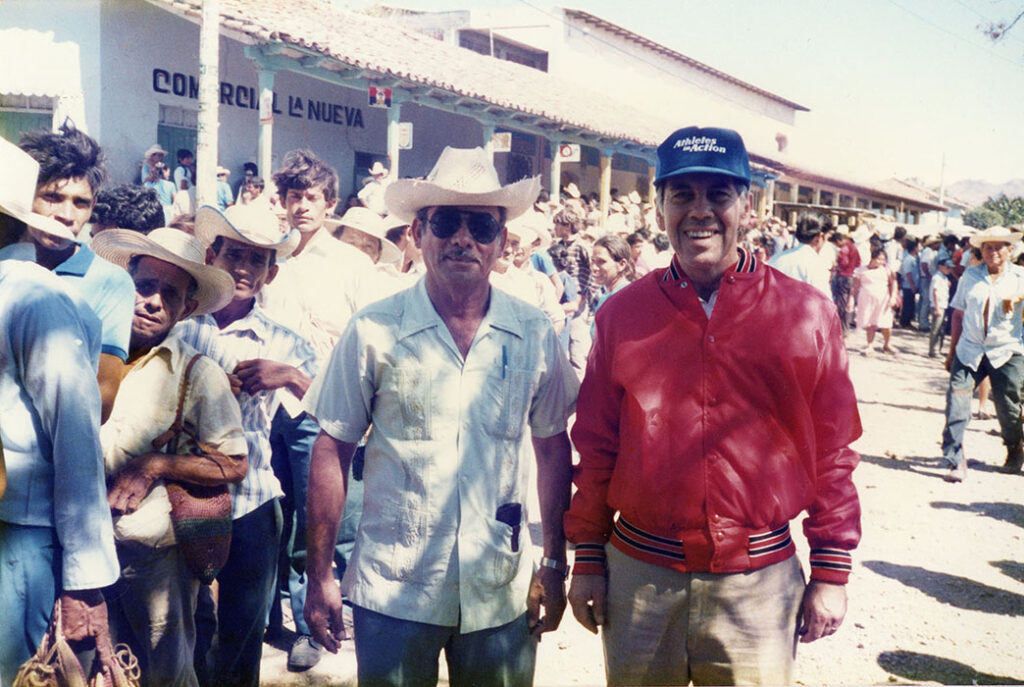 Photograph of an unknown man wearing a cowboy hat and sunglasses and Senator Richard Lugar. In the background, a long line of people waits to vote in the election.