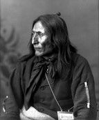 Black and white photo of Crowfoot, a Native American chief. Crowfoot is seated in semi-profile with the left-hand side of his face showing. He is wearing dark clothing and has chest-length hair.