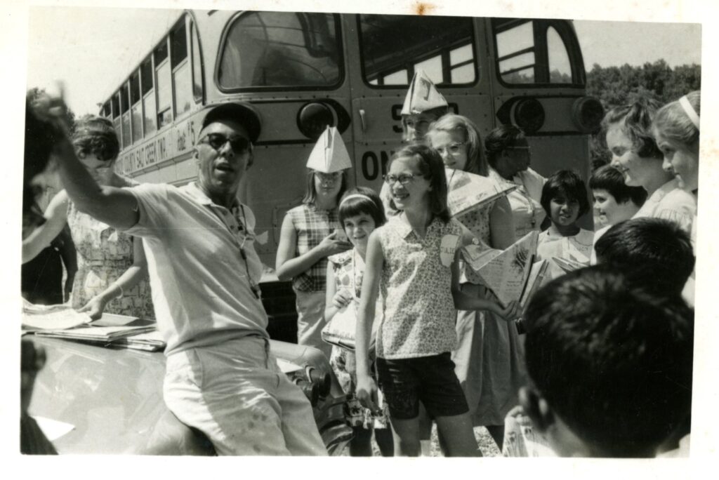 Black and white candid photo of Miller in front of a school bus with a group of children