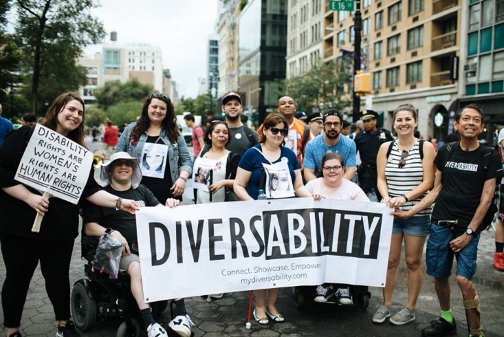 A group of chair users and people who are standing are holding a banner during a Disability Pride parade with tall buildings behind them. The banner says, “Diversability, Connect, Showcase, Empower, mydisability.com”. A woman to the left of banner holds a sign saying, “disability rights are women’s rights are human rights” with “human rights” double underscored. Three women standing behind the banner are wearing papers with their faces on them. 
