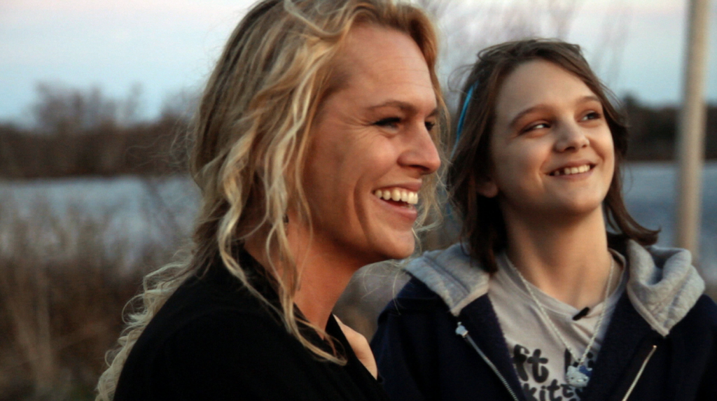 A film still from the documentary “Best and Most Beautiful Things” shows Michelle Smith, right, and her mother Julie, left, smiling at something off screen to the right. Behind them are weeds, a field, and trees. 