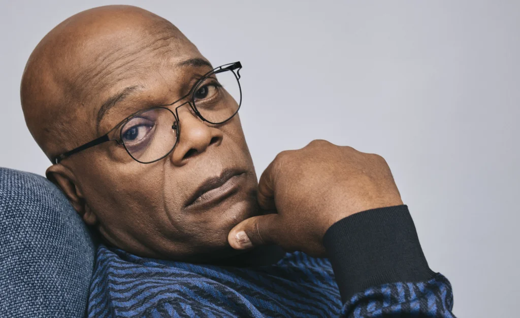 Samuel L Jackson is sitting on a couch staring at the camera while holding his hand on his chin.