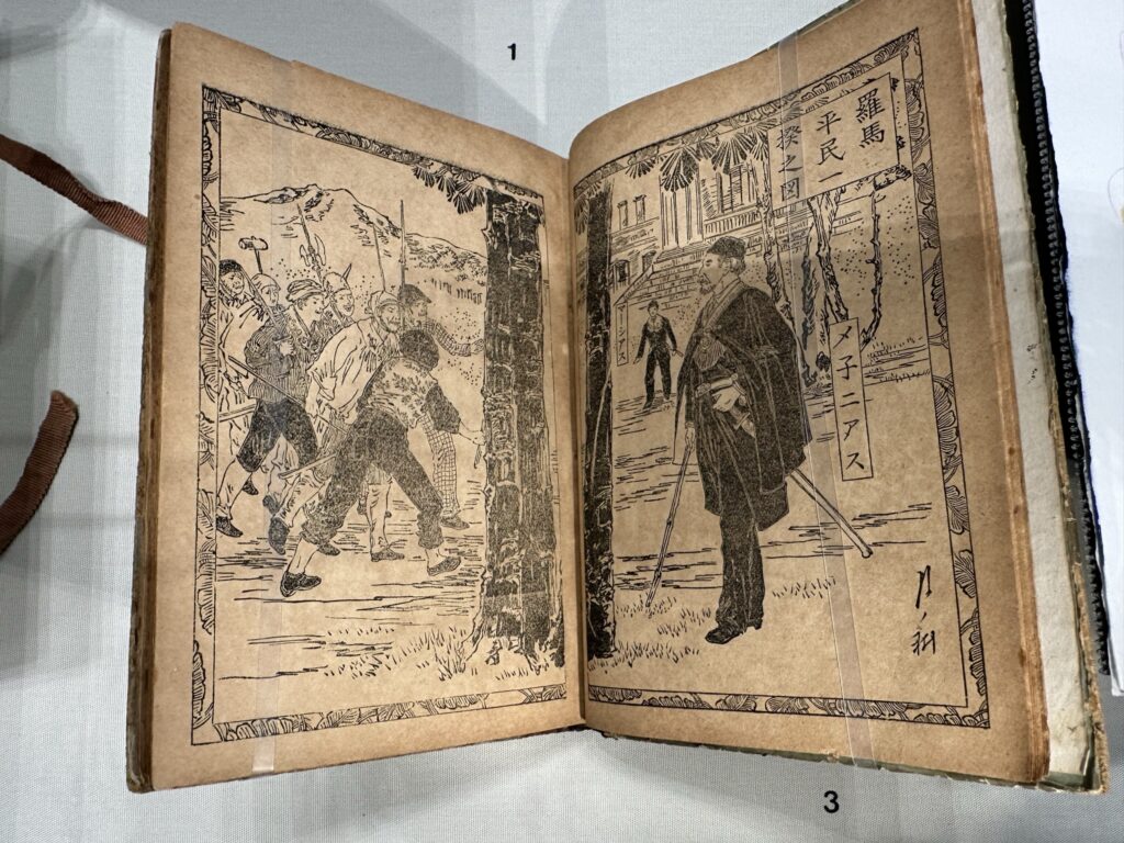 A book open to Japanese woodcuts of a scene from Coriolanus. Several men carry spears and axes while another man in a black cape with a sword at his side looks on.