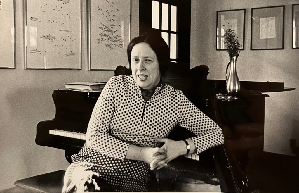 Portrait of Mary Ellen Solt sitting backwards on a piano bench, leaning on a piano, in a room with framed versions of her concrete poems hanging on the walls.