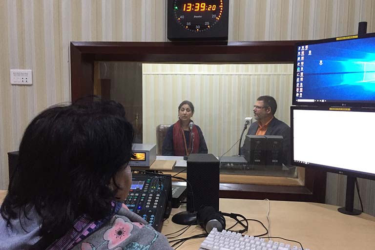 Rahat Bano Multanikar interviewing Alan Burdette in a studio at Radio Pakistan Multan while a technician sits at a desk with a computer workstation, speakers, and an audio control panel outside the studio’s glass window.