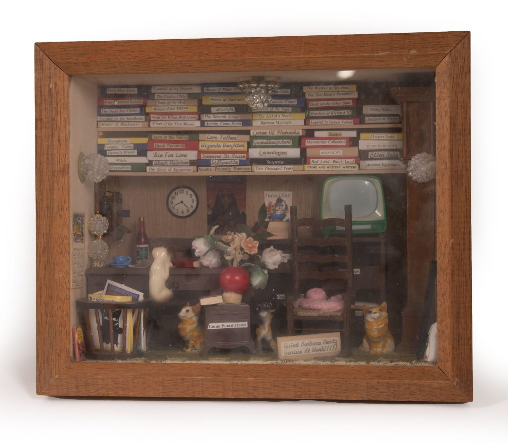 A diorama of an office in a wooden box with miniatures of Mertz’s works on a shelf above. Three ceramic miniature cats sit on the green carpeted floor next to a wooden basket of periodicals and a fourth cat climbs onto a desk with a teacup and a loose book. A sign at the bottom reads “Quiet Barbara Mertz Genius At Work!!!!”