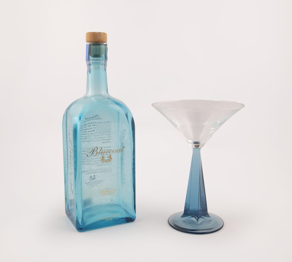 Blue translucent empty bottle of Bluecoat dry gin sits next to a martini glass with a blue translucent pyramid-like stem