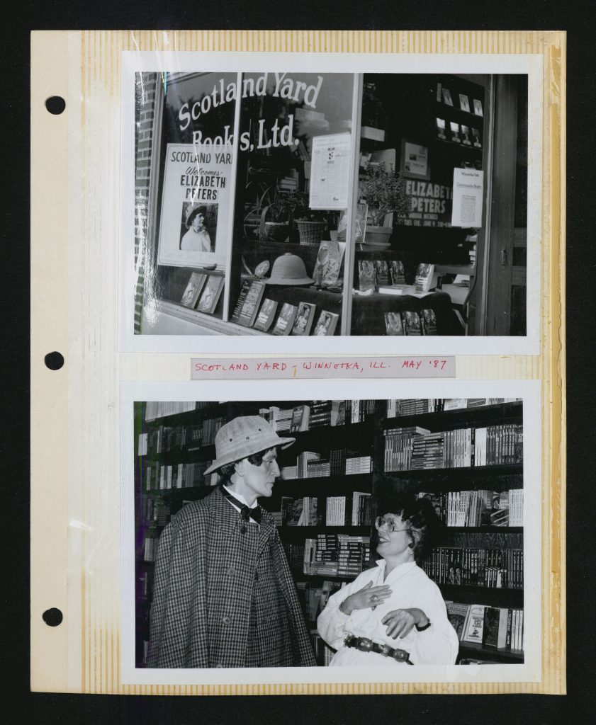 Page from a photo album labeled “Scotland Yard – Winnetka, Ill. May ‘87” in red ink in the middle of the page. Above photo is black and white view into a bookstore window where signs advertising a visit from Elizabeth Peters are prominently featured along with a pith helmet and many copies of Peters’s books. Below photo is black and white shot of Mertz smiling and pretending to swoon while looking at a wax figure of Sherlock Holmes