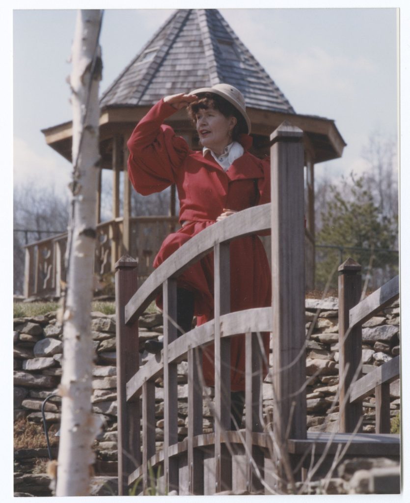 Mertz, dressed in a red Victorian suit and pith helmet, stands on a small bridge in front of a gazebo with one foot up on the wooden railing and her hand against her forehead in a surveying fashion, looking off into the distance