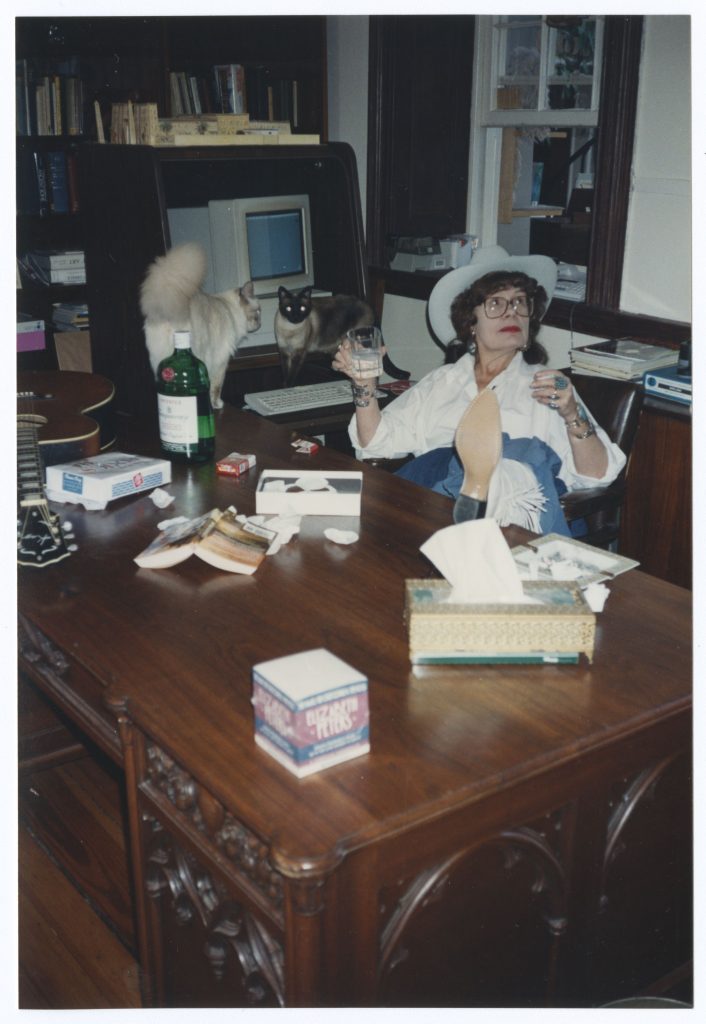 Mertz sits at a writing desk with her foot up on the table while smoking and looking away, a glass of gin in her hand. Two cats stand in front of a computer in the background
