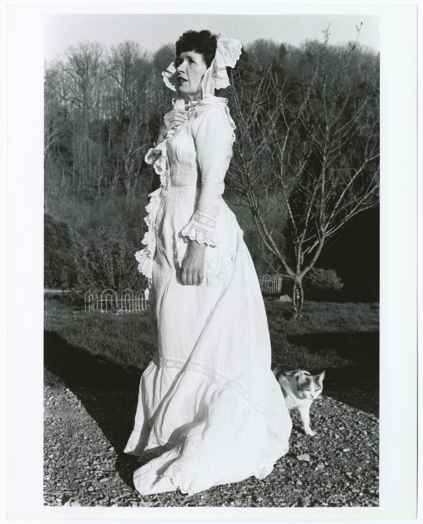 Mertz, dressed in a white lace dress with a bonnet, looks off into the distance while standing outside. A spotted cat walks behind her