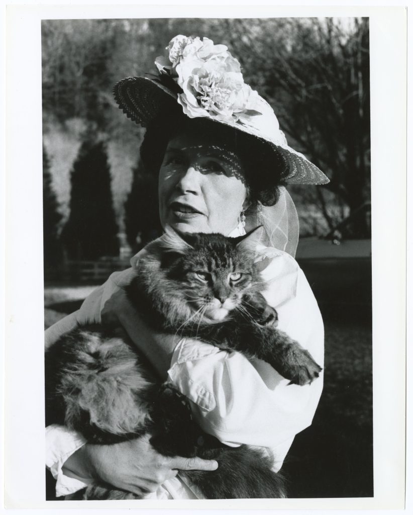 Mertz, wearing a straw hat with a large flower adornment, looks at the camera while holding a fluffy cat in both hands