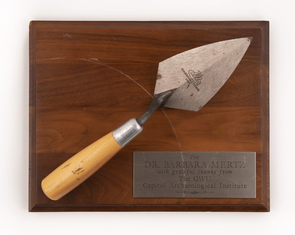 A wooden plaque with a trowel attached and a metal placard with the following text engraved: “For Dr. Barbara Mertz with grateful thanks from the GWU Capitol Archaeological Institute”