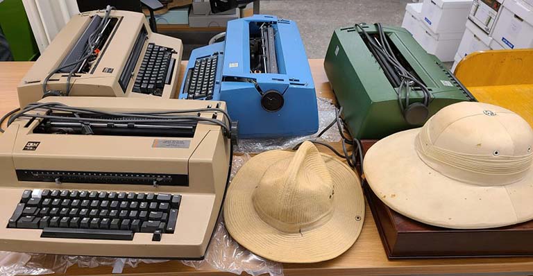 Four IBM typewriters (two tan, one blue, one green) sit on a large table next to a pith helmet and a khaki hat