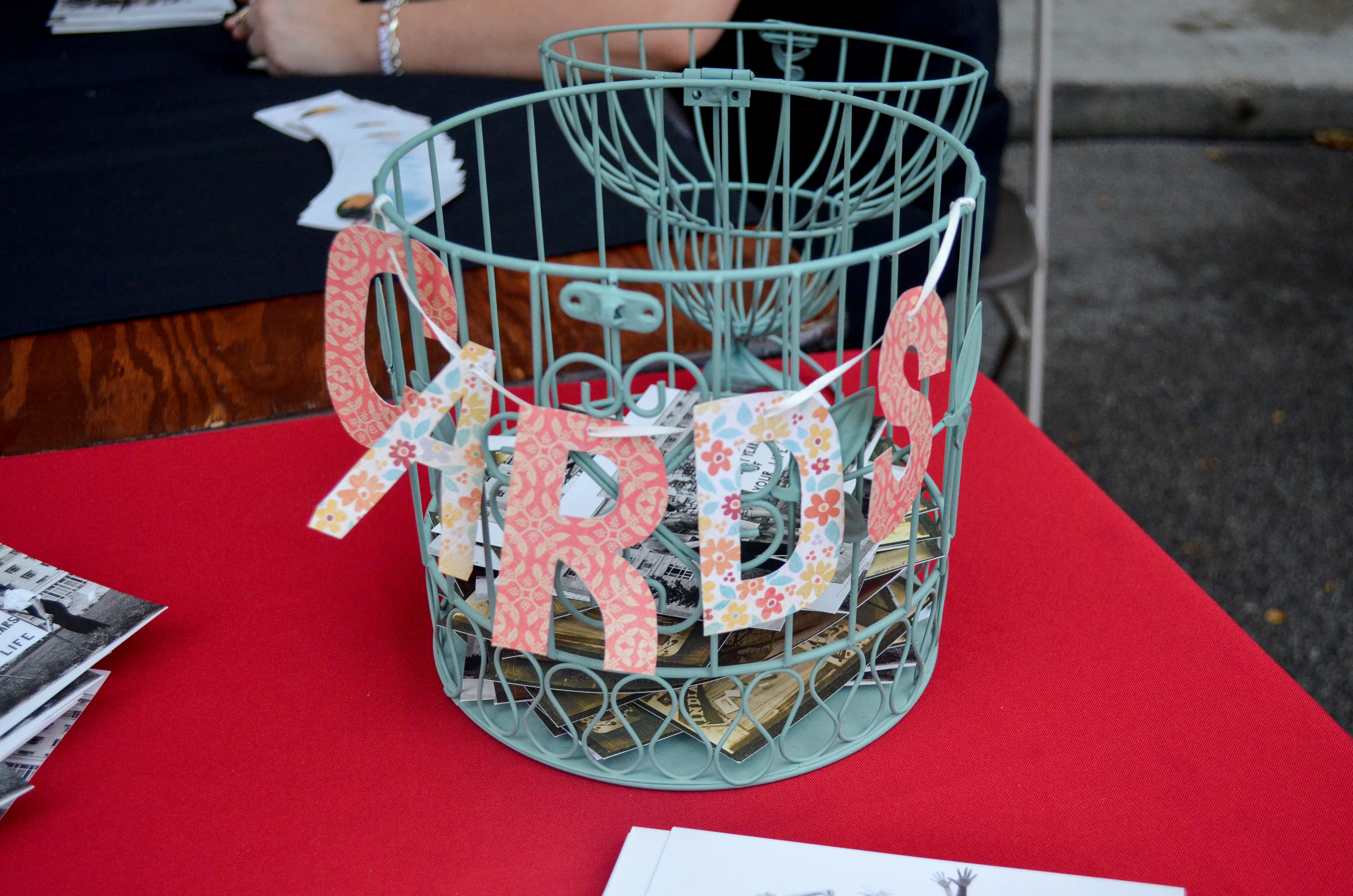 A decorative birdcage with a cut-out sign reading "CARDS" sits on a table. The birdcage is full of postcards.
