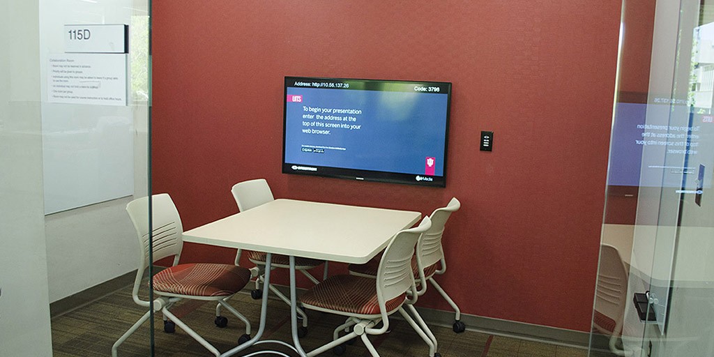Color photograph of a small meeting room with a table and four chairs and a monitor.