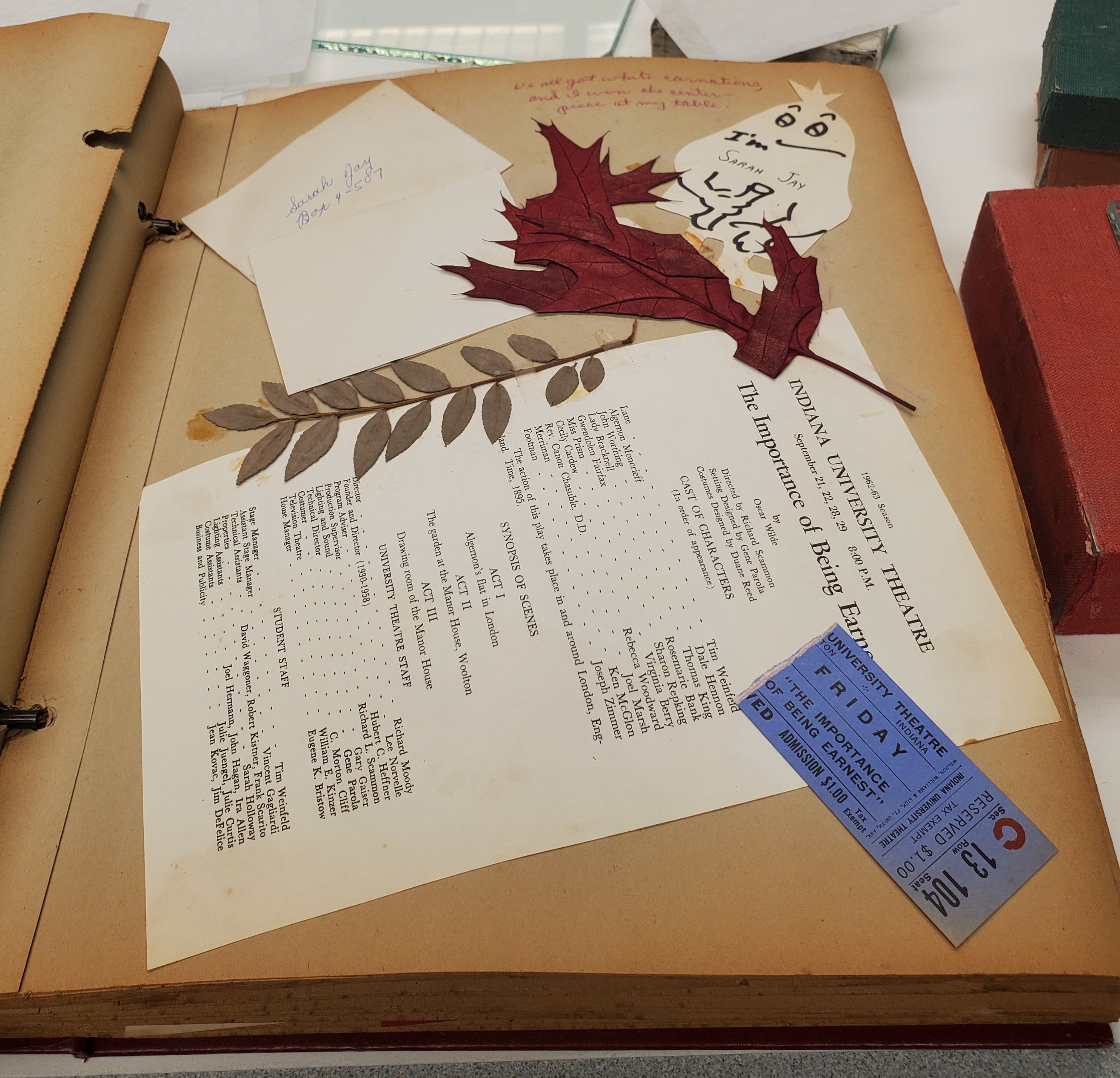 Scrapbook open to page containing a ticket stub, a theater program from the Importance of Being Earnest from the 1962-63 Indiana University Theatre Season, a crimson-colored leaf and a branch with many small leaves