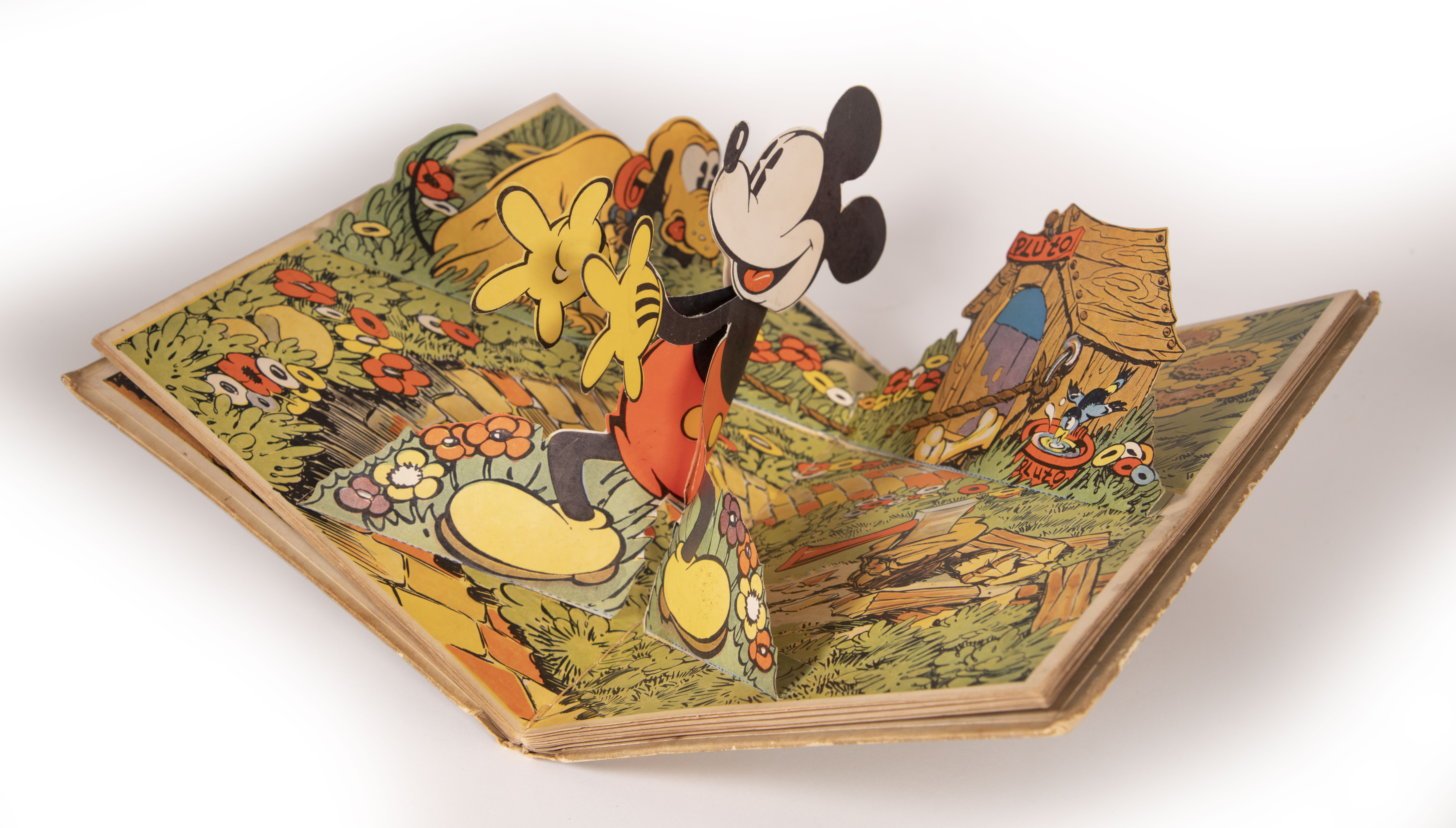 This photo of the book Pop-Up Mickey Mouse features a pop-up of Mickey standing up in his garden with his two arms pointed straight. Meanwhile, a pop-up of Pluto is tied to his doghouse looking perplexed.