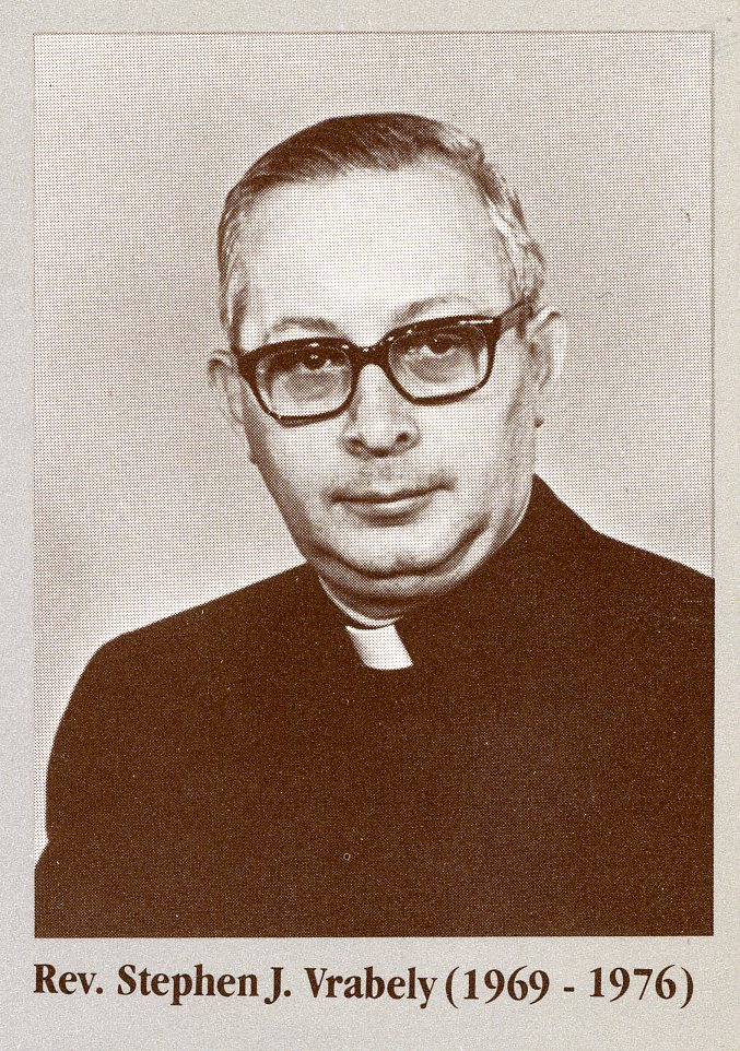 Black and white portrait showing Reverend Stephen J. Vrabely from the waste up smiling with his mouth closed. He is wearing glasses, a clerical collar, and a dark shirt. His skin tone is pale and his short dark hair is gray at the temples. He is positioned in front of a neutral background. His name is typed below the image followed by the years "1969 - 1976" typed in parentheses.